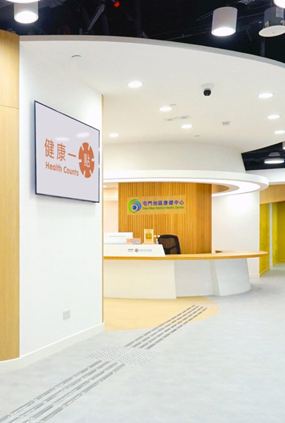 Reception area of the Tuen Mun District Health Centre (TMDHC) in Hong Kong, featuring a curved white desk, a wooden backdrop, and a 'Health Counts' digital sign